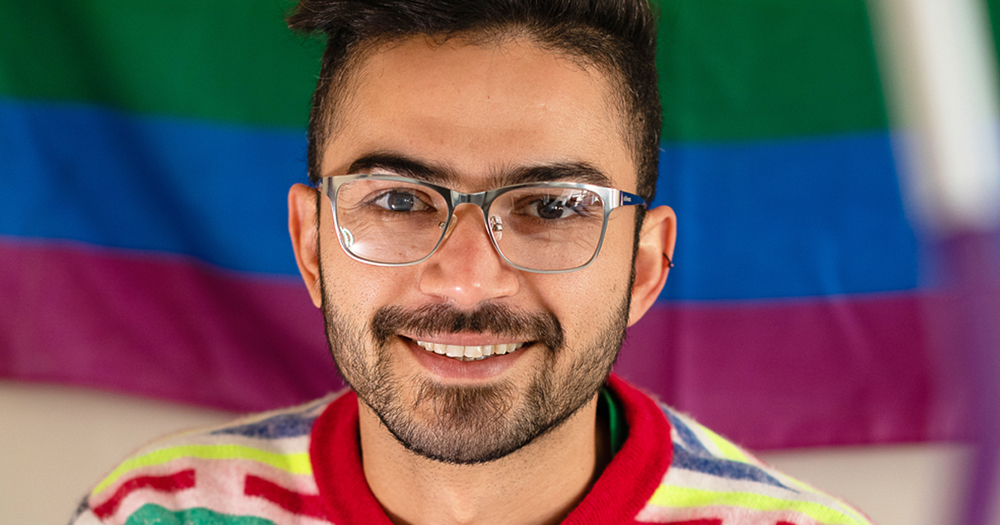 A young man with glasses smiling, behind him a Pride flag. Diego Caixeta from MPOWER talks about addressing the shame around sex.