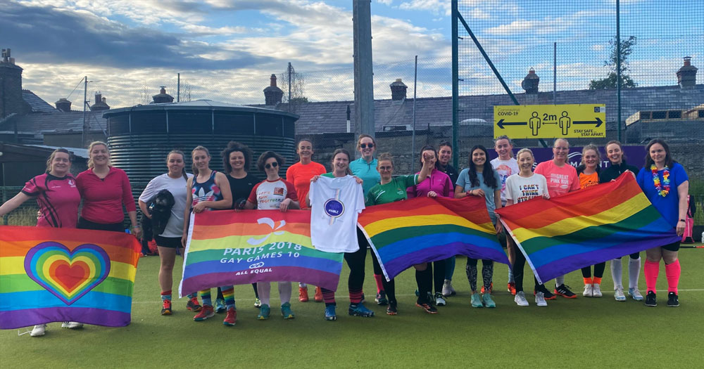 Photo of the Pink Ladies Hockey club standing on a pitch and waving rainbow flags